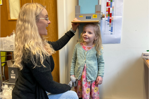 A woman crouched by a measure attached to the wall, measuring her daughter.